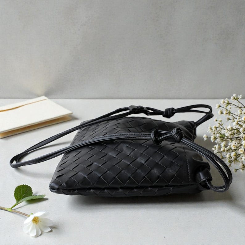 Puede incluir: Un bolso bandolera negro de cuero tejido con cierre de cord&oacute;n. El bolso tiene una correa larga y delgada y una forma rectangular. El bolso est&aacute; hecho de un material tejido y tiene una textura suave y flexible.