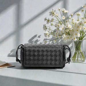 Puede incluir: Bolso bandolera de cuero tejido gris oscuro de forma rectangular. El bolso tiene cierre de solapa y asas negras. Un jarr&oacute;n con flores blancas en el fondo.
