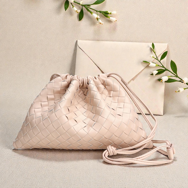 Puede incluir: Un bolso con cord&oacute;n de cuero tejido rosa claro con una correa para el hombro. La bolsa est&aacute; junto a un sobre a juego. Flores blancas y hojas verdes en el fondo.