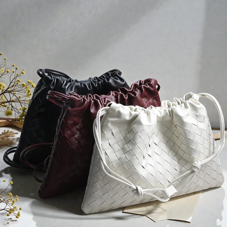 Puede incluir: Tres bolsos con cord&oacute;n de cuero tejido en negro, burdeos y blanco. Los bolsos tienen un patr&oacute;n de tejido de cesta y una parte superior fruncida con cierre de cord&oacute;n. El bolso blanco est&aacute; en primer plano, con los bolsos negro y burdeos detr&aacute;s.