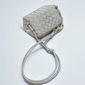 Pu&ograve; includere: Una borsa a tracolla in pelle intrecciata grigio chiaro con cerniera dorata e una tracolla lunga e sottile. La borsa ha una forma rettangolare con bordi arrotondati e un dettaglio annodato.