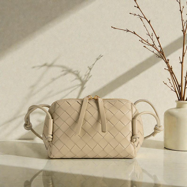 Pu&ograve; includere: Borsa a tracolla in pelle intrecciata beige con cerniera dorata e manici annodati. La borsa &egrave; rettangolare con un motivo intrecciato e si trova su una superficie bianca. Un vaso con rami &egrave; sullo sfondo.