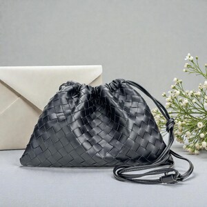 Puede incluir: Un bolso negro de cuero tejido con cord&oacute;n, correa larga y detalle de nudo. El bolso tiene forma triangular y un aspecto texturizado. Un sobre blanco y flores blancas se encuentran en el fondo.