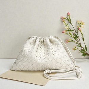 Puede incluir: Un bolso de hombro blanco de cuero tejido con cierre de cord&oacute;n. El bolso tiene una correa larga y delgada y una forma rectangular. El bolso est&aacute; sobre una superficie marr&oacute;n claro con flores rosas en el fondo.
