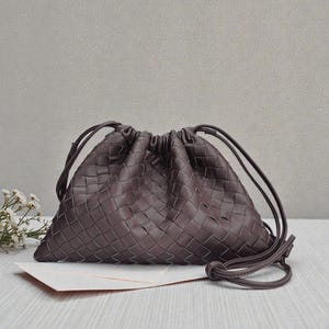 Puede incluir: Un bolso con cord&oacute;n tejido de color marr&oacute;n oscuro con forma triangular. El bolso presenta un patr&oacute;n trenzado y un cierre con cord&oacute;n. El bolso est&aacute; sobre una superficie blanca con unas peque&ntilde;as flores blancas en el fondo.