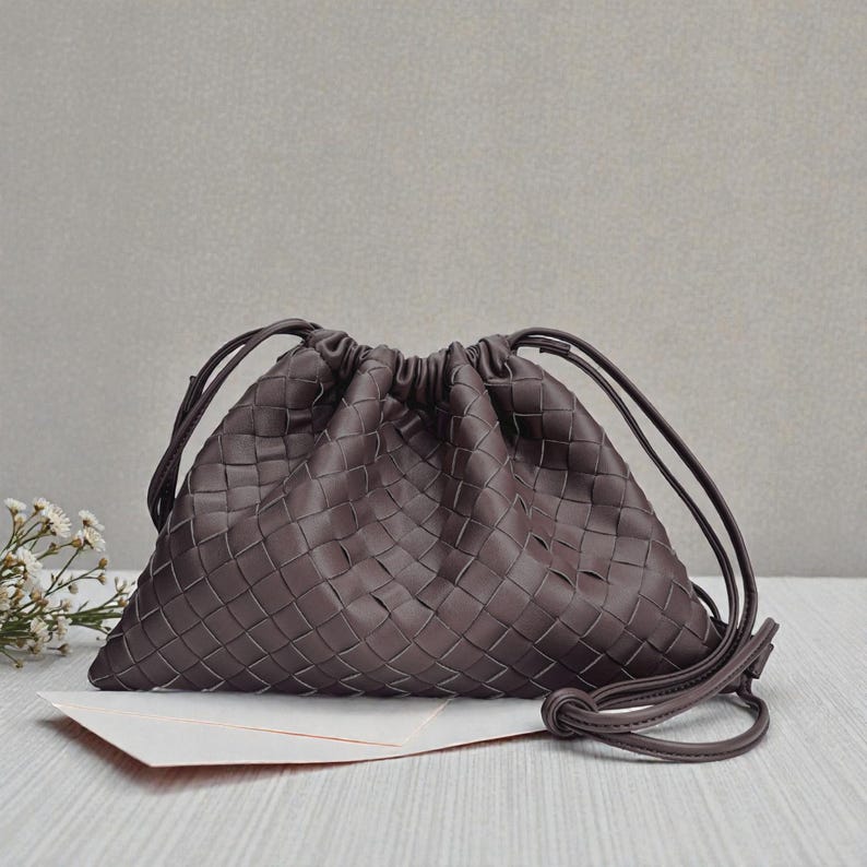 Puede incluir: Un bolso con cord&oacute;n tejido de color marr&oacute;n oscuro con forma triangular. El bolso presenta un patr&oacute;n trenzado y un cierre con cord&oacute;n. El bolso est&aacute; sobre una superficie blanca con unas peque&ntilde;as flores blancas en el fondo.
