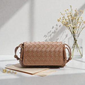 Puede incluir: Un bolso cruzado de cuero tejido de color marr&oacute;n claro con forma rectangular. El bolso presenta un cierre de solapa y detalles de correas anudadas. El bolso se exhibe sobre una superficie blanca con un jarr&oacute;n de flores en el fondo.