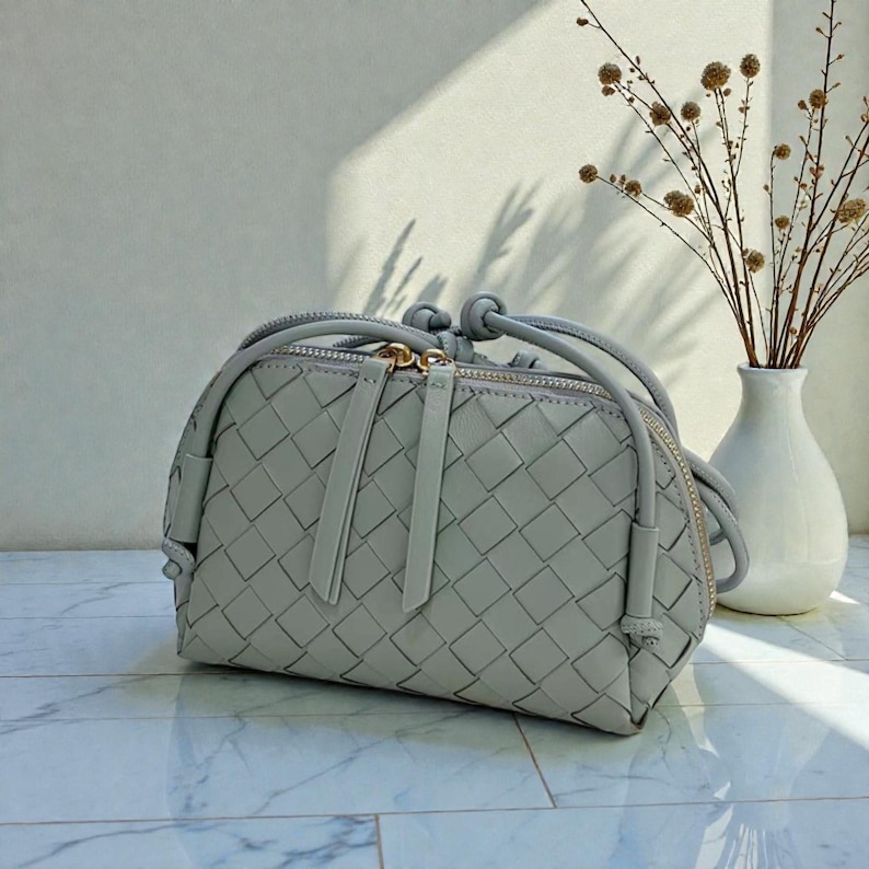 Pu&ograve; includere: Una pochette in pelle intrecciata grigio chiaro, di forma arrotondata, con cerniera dorata. La borsa presenta un cinturino annodato e due lunghe tirette. Un vaso bianco con fiori secchi &egrave; sullo sfondo.