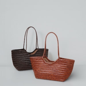 May include: Two woven tote bags. One is dark brown, the other is a lighter reddish-brown. Both have a rectangular shape with rounded corners and a woven pattern. The handles are braided and arched. The bags are empty.