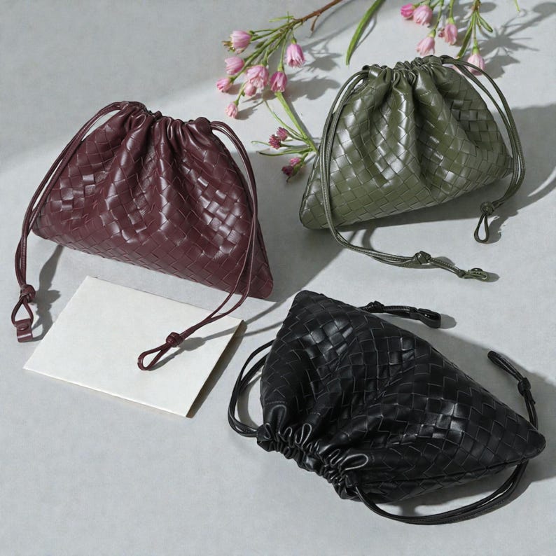 Puede incluir: Tres bolsas con cord&oacute;n de cuero tejido en burdeos, verde oliva y negro. Cada bolsa tiene una parte superior fruncida con un cierre de cord&oacute;n. Las bolsas est&aacute;n dispuestas sobre una superficie gris claro con una ramita de flores rosas.