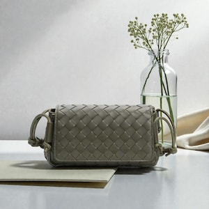 Puede incluir: Un peque&ntilde;o bolso tejido de color verde oliva, de forma rectangular y con cierre de solapa. El bolso tiene una correa para el hombro y se exhibe sobre una superficie blanca. Un jarr&oacute;n de cristal transparente con delicadas flores blancas est&aacute; en el fondo.