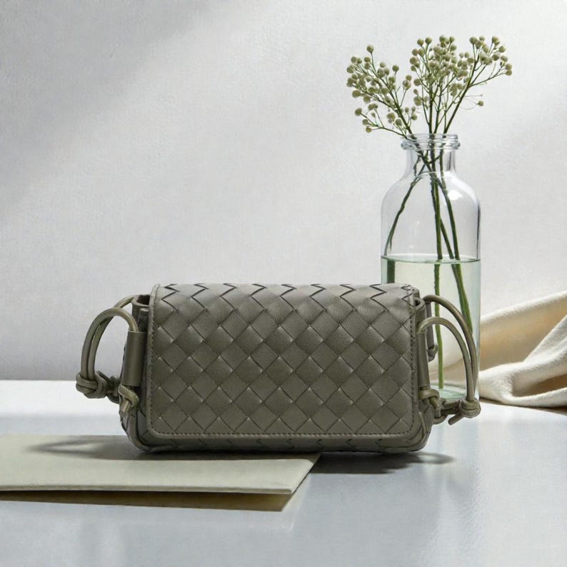 Puede incluir: Un peque&ntilde;o bolso tejido de color verde oliva, de forma rectangular y con cierre de solapa. El bolso tiene una correa para el hombro y se exhibe sobre una superficie blanca. Un jarr&oacute;n de cristal transparente con delicadas flores blancas est&aacute; en el fondo.
