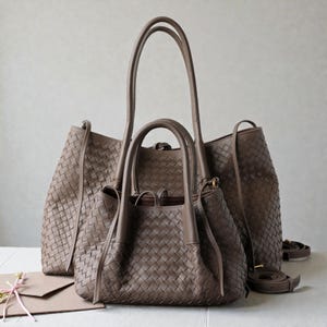 Peut inclure: Trois sacs à main en cuir tressé marron de différentes tailles. Le plus grand a de longues poignées et une bandoulière. Le sac du milieu a des poignées plus courtes. Le plus petit sac a une fermeture à cordon. Les sacs sont sur une surface blanche.