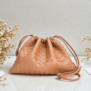 Puede incluir: Un bolso con cord&oacute;n de cuero tejido de color marr&oacute;n claro. El bolso tiene forma triangular con la parte superior fruncida y correas largas y finas. El patr&oacute;n tejido es visible en toda la superficie del bolso.