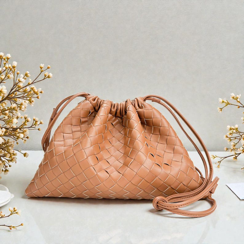 Puede incluir: Un bolso con cord&oacute;n de cuero tejido de color marr&oacute;n claro. El bolso tiene forma triangular con la parte superior fruncida y correas largas y finas. El patr&oacute;n tejido es visible en toda la superficie del bolso.
