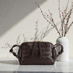 Pu&ograve; includere: Una borsa a tracolla in pelle intrecciata marrone scuro con cerniera dorata e manici annodati. La borsa &egrave; rettangolare con la parte superiore arrotondata. Un vaso bianco con una composizione floreale &egrave; sullo sfondo.