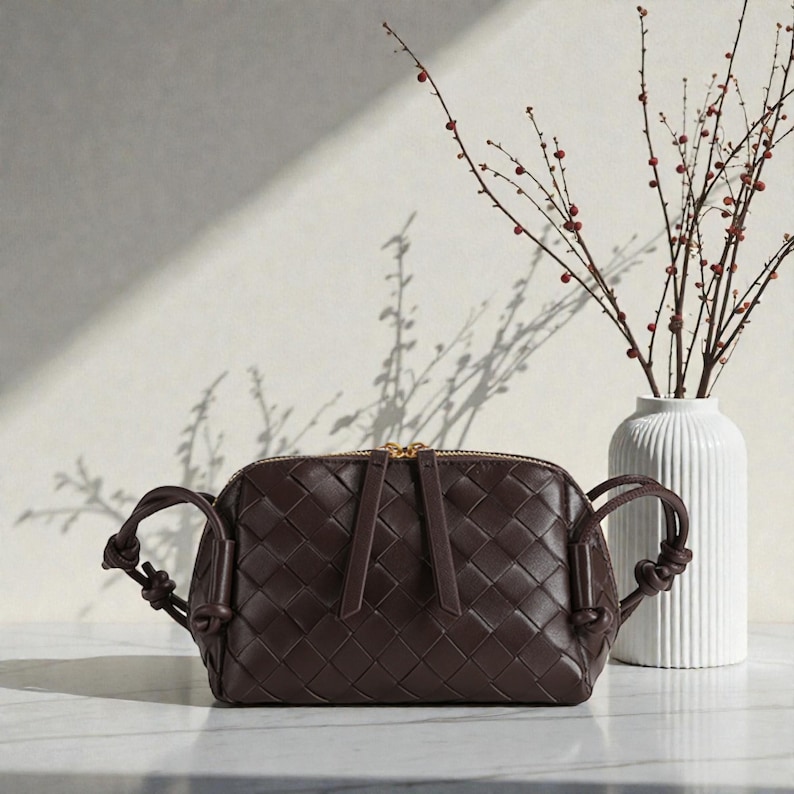Pu&ograve; includere: Una borsa a tracolla in pelle intrecciata marrone scuro con cerniera dorata e manici annodati. La borsa &egrave; rettangolare con la parte superiore arrotondata. Un vaso bianco con una composizione floreale &egrave; sullo sfondo.