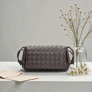 Puede incluir: Un bolso bandolera de cuero tejido de color marr&oacute;n oscuro. El bolso tiene forma rectangular con bordes redondeados y asas anudadas. Un jarr&oacute;n de cristal transparente con delicadas flores y ramitas blancas est&aacute; detr&aacute;s del bolso.