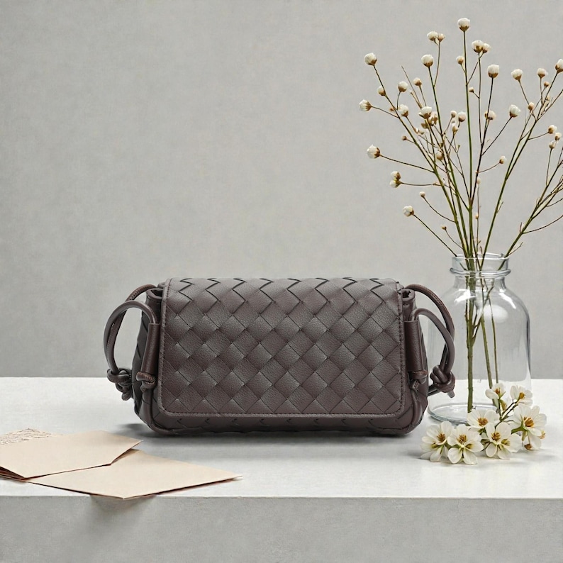 Puede incluir: Un bolso bandolera de cuero tejido de color marr&oacute;n oscuro. El bolso tiene forma rectangular con bordes redondeados y asas anudadas. Un jarr&oacute;n de cristal transparente con delicadas flores y ramitas blancas est&aacute; detr&aacute;s del bolso.