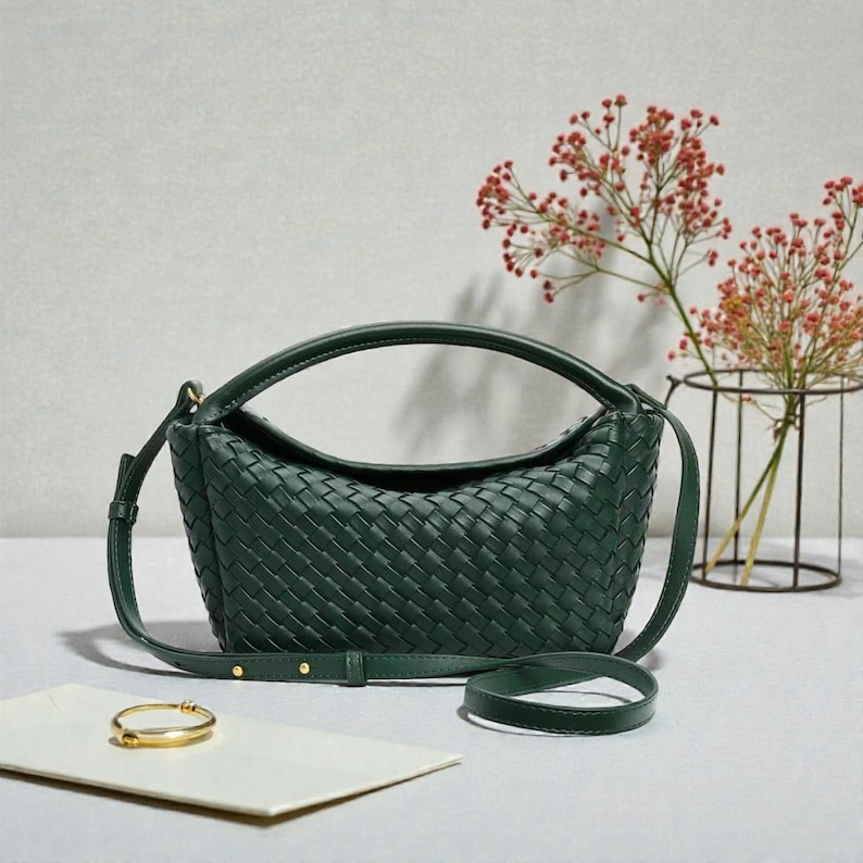 May include: A dark green woven handbag with a curved top handle and a detachable shoulder strap. A gold bracelet rests on a white card. Red flowers in a black wire vase are in the background.