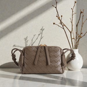 Pu&ograve; includere: Una borsa a tracolla in pelle intrecciata color talpa con chiusura a zip e manici annodati. La borsa &egrave; di forma ovale e poggia su una superficie bianca. Un vaso bianco con rami &egrave; sullo sfondo.
