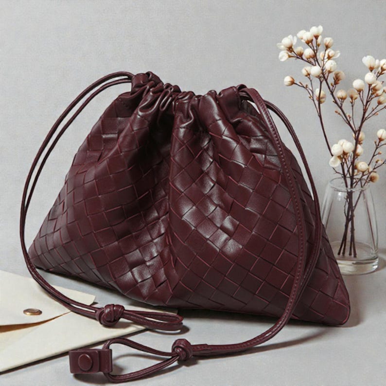 Puede incluir: Bolso con cord&oacute;n de cuero burdeos con un patr&oacute;n tejido. El bolso tiene un cierre de cord&oacute;n anudado y un peque&ntilde;o cierre rectangular. Se ve un peque&ntilde;o sobre blanco con un bot&oacute;n dorado.