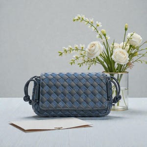 Puede incluir: Un bolso cruzado azul y gris tejido con cierre de solapa. El bolso tiene un dise&ntilde;o trenzado y una correa anudada. Un jarr&oacute;n con rosas blancas y una tarjeta en blanco est&aacute;n en el fondo.
