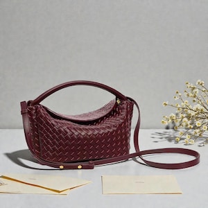 May include: A burgundy woven leather handbag with a curved handle and a long, adjustable shoulder strap. The bag has a flap closure and gold-toned hardware. The bag is approximately 30 cm wide and 20 cm tall.