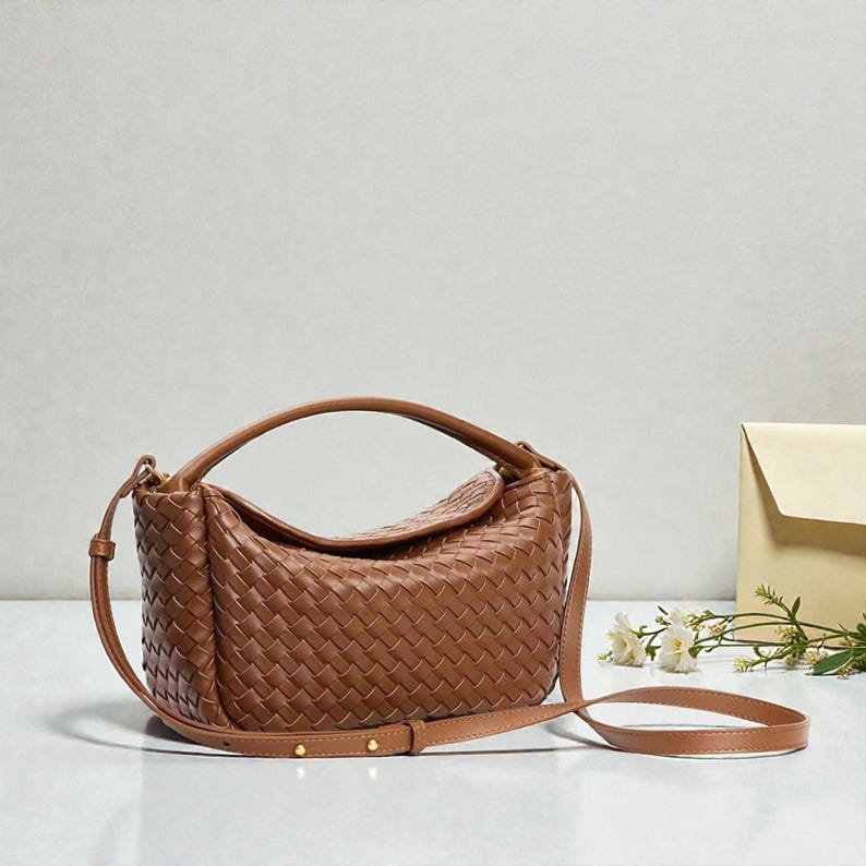 May include: A brown woven leather handbag with a top handle and a detachable shoulder strap. The bag has a curved shape and a gold-toned zipper. The bag is approximately 30 cm wide.