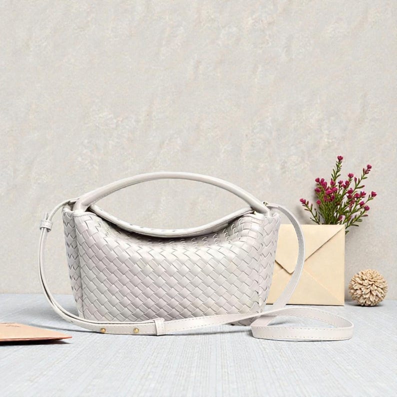 May include: A pale grey woven leather handbag with a top handle and a detachable shoulder strap. The bag is rectangular with rounded edges. The background includes a small vase with pink flowers, an envelope, and a decorative object.