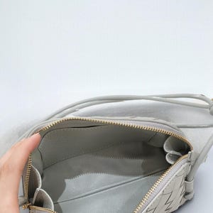 Pu&ograve; includere: Una borsa a tracolla in pelle intrecciata grigio chiaro con cerniera dorata. La borsa &egrave; aperta, rivelando un interno grigio. Anche la tracolla &egrave; grigia. La borsa &egrave; di forma rettangolare.