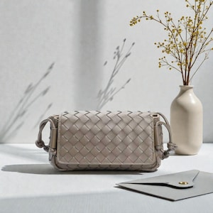 Puede incluir: Un bolso bandolera de cuero tejido gris claro con cierre de solapa. El bolso tiene forma rectangular con esquinas redondeadas y una correa anudada. Un peque&ntilde;o sobre gris con un bot&oacute;n dorado est&aacute; junto al bolso. Un jarr&oacute;n blanco con flores amarillas est&aacute; en el fondo.