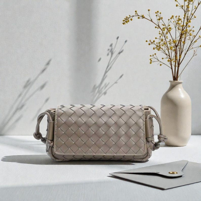 Puede incluir: Un bolso bandolera de cuero tejido gris claro con cierre de solapa. El bolso tiene forma rectangular con esquinas redondeadas y una correa anudada. Un peque&ntilde;o sobre gris con un bot&oacute;n dorado est&aacute; junto al bolso. Un jarr&oacute;n blanco con flores amarillas est&aacute; en el fondo.