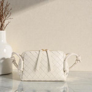 Pu&ograve; includere: Borsa a tracolla in pelle intrecciata bianco sporco, di forma rettangolare e con cerniera dorata. La borsa ha manici annodati su ogni lato ed &egrave; esposta su una superficie di marmo. Un vaso bianco con rami secchi &egrave; sullo sfondo.