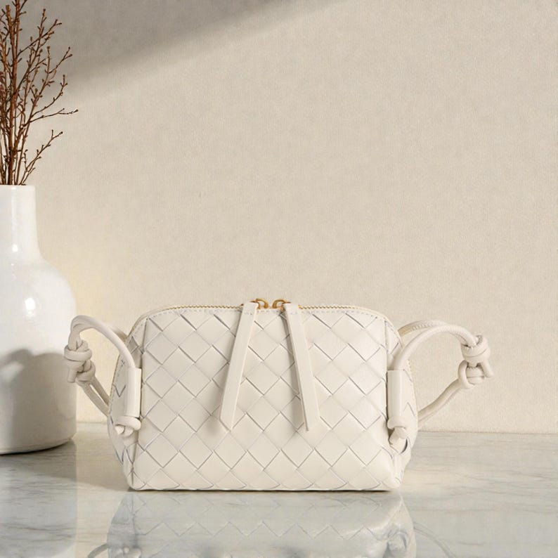 Pu&ograve; includere: Borsa a tracolla in pelle intrecciata bianco sporco, di forma rettangolare e con cerniera dorata. La borsa ha manici annodati su ogni lato ed &egrave; esposta su una superficie di marmo. Un vaso bianco con rami secchi &egrave; sullo sfondo.