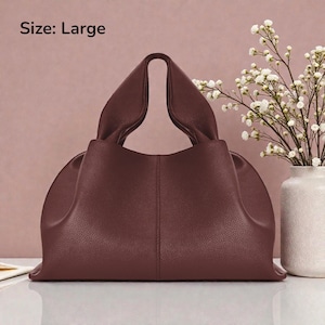 May include: A large, burgundy-colored handbag with a unique, gathered handle design. The bag has a structured shape and a smooth, leather-like texture. The text "Size: Large" is visible in the upper left corner. A vase of white flowers is in the background.