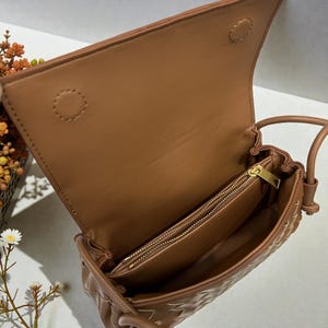 Puede incluir: Bolso de mano de cuero marr&oacute;n claro con un dise&ntilde;o tejido, abierto para mostrar el interior. El bolso tiene un cierre de solapa y un bolsillo interior con cremallera. El bolso es de color marr&oacute;n claro y tiene una correa para el hombro.