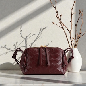 Pu&ograve; includere: Borsa a tracolla in pelle bordeaux con design intrecciato. La borsa ha una cerniera dorata e manici annodati. Un vaso bianco con rami &egrave; a destra della borsa.