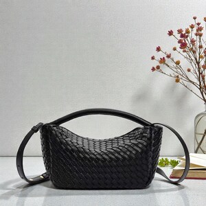 May include: A black woven leather handbag with a curved top handle and a detachable shoulder strap. The bag has a rectangular shape with a woven pattern. The bag is sitting on a light grey surface.