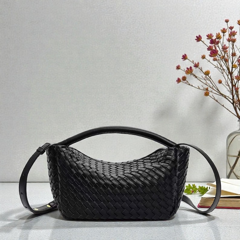 May include: A black woven leather handbag with a curved top handle and a detachable shoulder strap. The bag has a rectangular shape with a woven pattern. The bag is sitting on a light grey surface.
