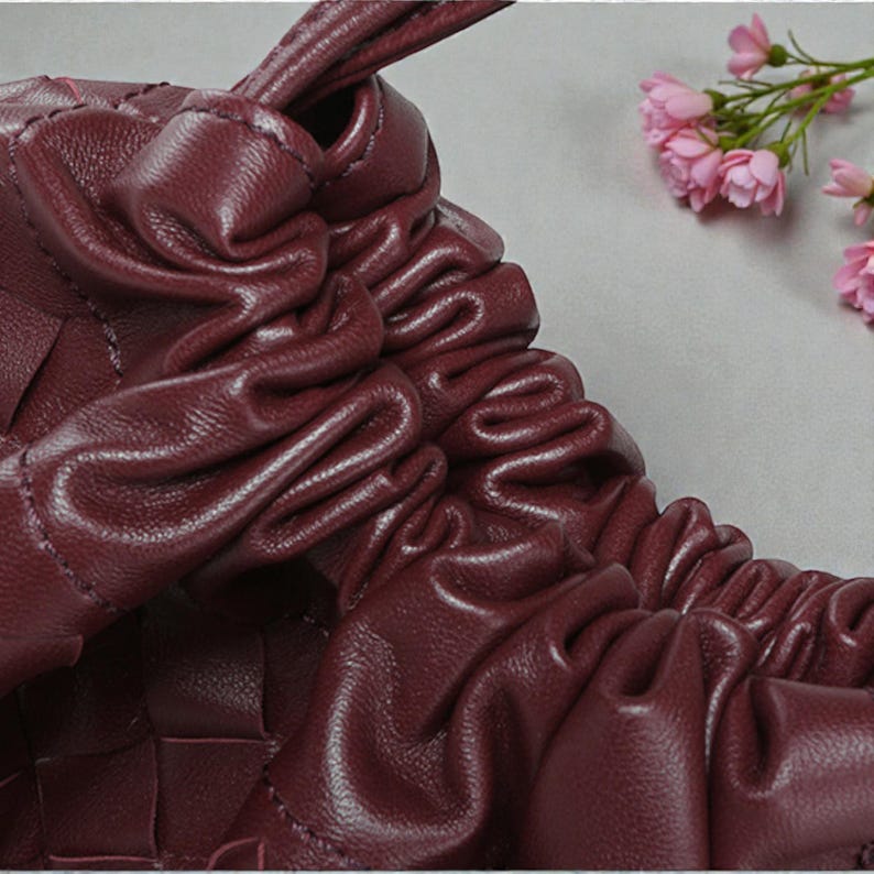Puede incluir: Primer plano de un bolso de mano de cuero burdeos con un dise&ntilde;o tejido. El bolso presenta detalles fruncidos y un asa anudada. Se ven flores rosas en el fondo, a&ntilde;adiendo un toque de color a la imagen.