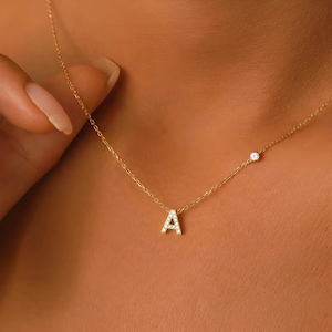 Könnte beinhalten: Eine zarte goldene Halskette mit einem Anhänger in Form des Buchstabens "A", besetzt mit Diamanten. Die Halskette enthält auch einen kleinen, runden Diamanten. Die Kette ist fein und das Design elegant und minimalistisch.