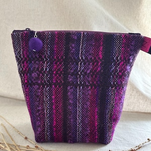 May include: A purple and pink woven fabric pouch with a zipper closure and a small purple pom-pom attached. The pouch has a rectangular shape with a slight trapezoid design. The fabric has vertical stripes of varying shades of purple and pink.