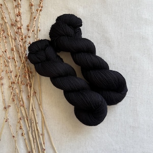 May include: Two skeins of black yarn are displayed on a neutral-toned fabric. The yarn is tightly twisted, showcasing its texture and depth. Dried branches with small buds are arranged to the left of the yarn, adding a natural element to the composition.