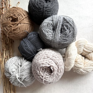 May include: A collection of seven yarn balls in various neutral shades including charcoal, light gray, beige, cream, and dark brown, arranged alongside a cluster of dried, thin, light-colored twigs with small buds on a textured fabric surface.