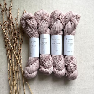 May include: Four skeins of dusty rose-colored yarn with white labels that read "les garcons SHIBUI KNITS HAVEN." The yarn is arranged on a neutral-toned fabric, with dried branches on the left side.