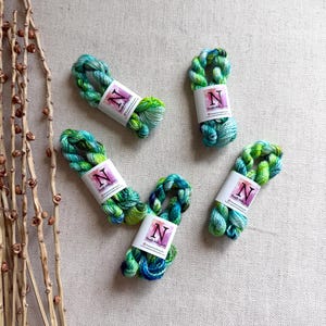 May include: Five skeins of hand-dyed yarn in shades of teal, green, and blue. Each skein is wrapped with a white label featuring the letter "N" in a purple square. The yarn is arranged on a neutral-colored surface.
