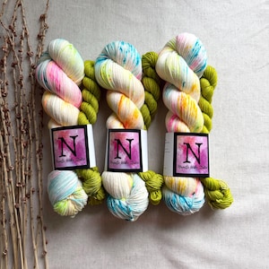 May include: Three skeins of colorful yarn with a white base, dyed with pink, yellow, blue, and orange hues. The yarn is paired with a bright green base. Each skein has a label with a large letter "N" and the text "Neville Wake Studio".