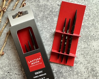 May include: A set of four black interchangeable knitting needles in a red and black box. The box has the text "Lantern Moon Handcrafted Ebony Cable Needles US 3, 6, 8 3.25, 4.0, 5.0 mm" printed on it.