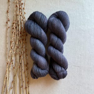 May include: Two skeins of dark charcoal gray yarn twisted into a classic hank shape. The yarn features a subtle heathered texture with visible fiber strands, presented alongside a cluster of thin, dried, light brown decorative twigs.